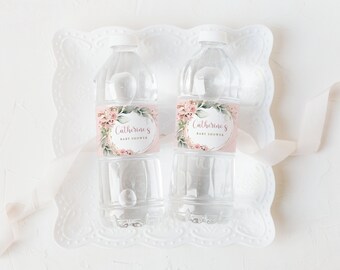 Könnte beinhalten: Zwei durchsichtige Plastikwasserflaschen mit rosa und goldenen floralen Etiketten, auf denen "Catherine's Baby Shower" steht. Die Flaschen befinden sich auf einer weißen Spitzentischdecke.