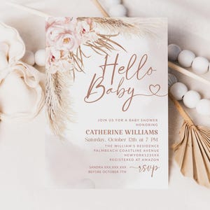 May include: A baby shower invitation with the words "Hello Baby" in a stylish script font. The invitation features floral and pampas grass accents in soft pink and beige tones. Details include the honoree's name, date, time, and location.