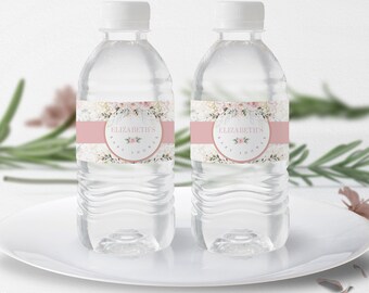 Könnte beinhalten: Zwei transparente Wasserflaschen mit weißen Verschlüssen. Jede Flasche hat ein rosa-blumiges Etikett mit der Aufschrift "ELIZABETH'S BABY SHOWER". Die Flaschen stehen auf einem weißen Teller, mit Grünzeug im Hintergrund.