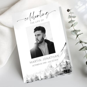 May include: A black and white photo memorial card with a black and white photo of a man. The card has a black and white watercolor background with a mountain scene. The text reads "celebrating the life of Martin Jonathan October 20, 1985 - July 13, 2018".