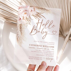 May include: A baby shower invitation with the words "Hello Baby" in elegant script. The invitation features floral and pampas grass accents in soft pink and beige tones. Details include the honoree's name, date, time, and location.
