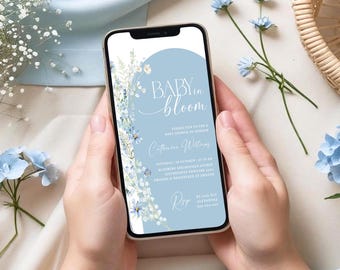 May include: A smartphone displays a baby shower invitation with the text "Baby in Bloom." The invitation features a light blue background with floral accents. The phone is held in a person's hands, with flowers in the background.