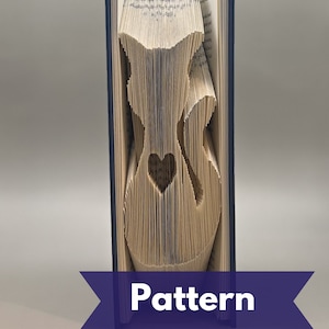 May include: A folded book with a heart-shaped cutout in the center. The pages are cut to form a silhouette of a cat. The book has a blue spine and the word "Pattern" is displayed on a purple banner.