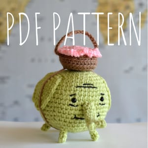 May include: A green crocheted amigurumi elephant with a brown crocheted basket on its head. The basket has a pink crocheted flower inside. The elephant has black eyes and a small brown crocheted trunk.
