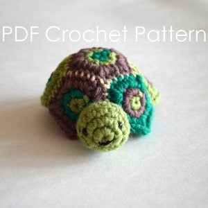 Puede incluir: Un juguete de amigurumi de tortuga tejido a crochet con una concha marrón, verde y turquesa. La tortuga tiene una cabeza verde con ojos negros y una pequeña sonrisa.
