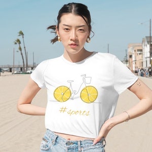 May include: White crop top with a graphic of a bicycle with lemon slices for wheels. The text '#sports' is printed below the bicycle.