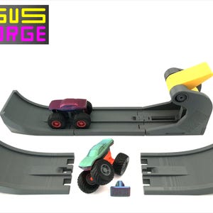 May include: A grey toy monster truck track set with two monster trucks. One truck is purple, the other is green. The track has a yellow launch mechanism and the text "GUS FORGE" in yellow and purple.