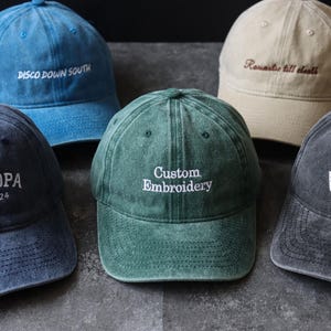 May include: Five baseball caps in various colors including blue, dark blue, green, beige, and charcoal gray. Each cap features embroidered text: DISCO DOWN SOUTH, GRANDPA EST. 2024, Custom Embroidery, Romantic till death, and FORUM in white or brown thread.