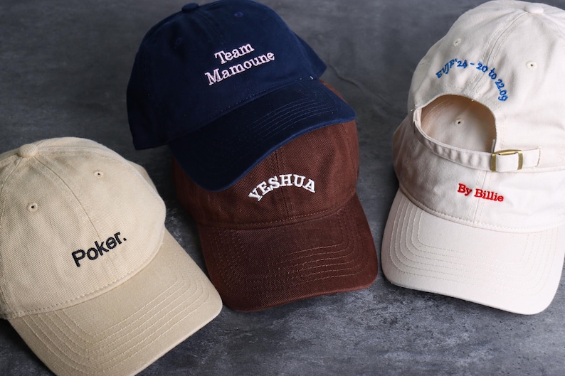 May include: Four baseball caps in various colors: navy blue, brown, and beige. Each cap has embroidered text: "Team Mamoune", "YESHUA", "Poker.", and "By Billie". The beige cap also has text on the back.