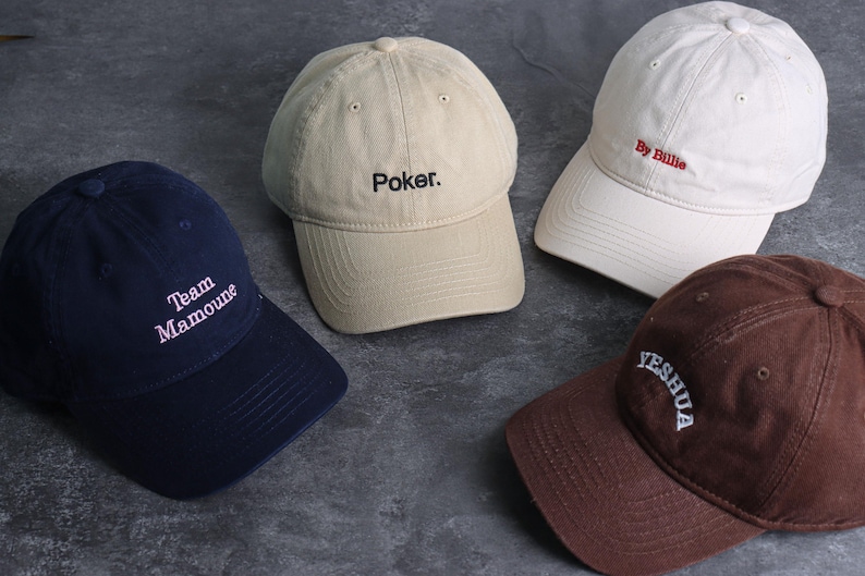 May include: Four baseball caps in navy, tan, white, and brown. Each cap has embroidered text: "Team Mamoune", "Poker", "By Billie", and "YESHUA". The caps are arranged on a dark gray surface.