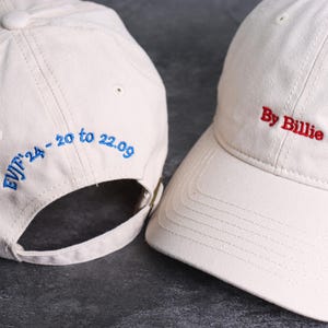 May include: Two off-white baseball caps. One cap has the text "By Billie" embroidered in red. The other cap has the text "EVJF'24 - 20 to 22.09" embroidered in blue. Both caps have a curved brim and adjustable strap.