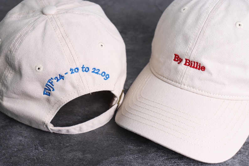 May include: Two off-white baseball caps. One cap has the text "By Billie" embroidered in red. The other cap has the text "EVJF'24 - 20 to 22.09" embroidered in blue. Both caps have a curved brim and adjustable strap.