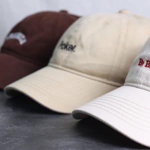 May include: Three baseball caps in a row, each with a curved brim and adjustable strap. The caps are in brown, tan, and white. The brown cap has white embroidered text, the tan cap has the word "Poker" embroidered in black, and the white cap has "By Billie" embroidered in red.