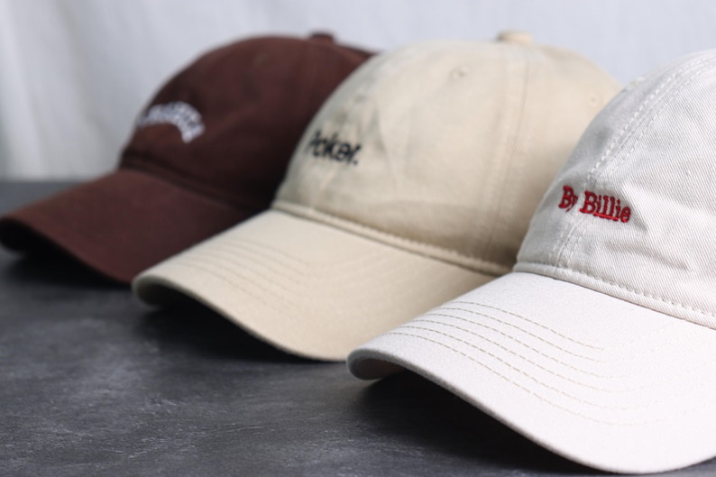 May include: Three baseball caps in a row, each with a curved brim and adjustable strap. The caps are in brown, tan, and white. The brown cap has white embroidered text, the tan cap has the word "Poker" embroidered in black, and the white cap has "By Billie" embroidered in red.