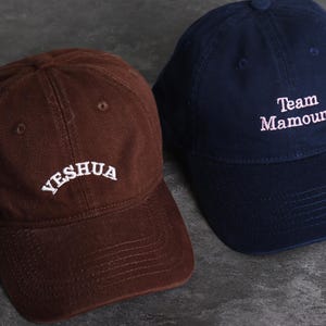 May include: Two baseball caps. One is brown with the word "YESHUA" embroidered in white. The other is navy blue with "Team Mamoune" embroidered in white. Both caps have a curved brim and a button on top.
