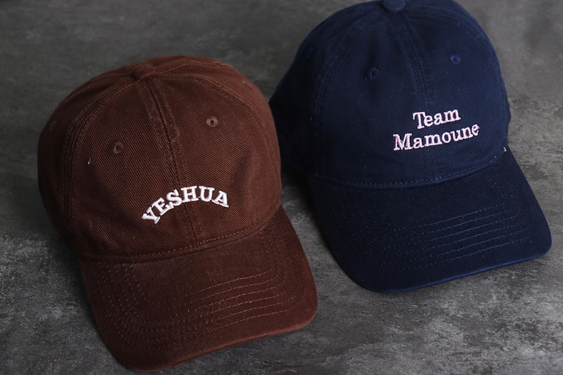 May include: Two baseball caps. One is brown with the word "YESHUA" embroidered in white. The other is navy blue with "Team Mamoune" embroidered in white. Both caps have a curved brim and a button on top.