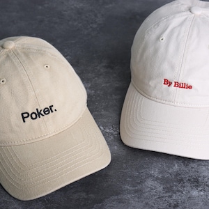 May include: Two baseball caps. One is tan with the word "Poker." embroidered in black. The other is white with "By Billie" embroidered in red. Both caps have a curved brim and adjustable strap.