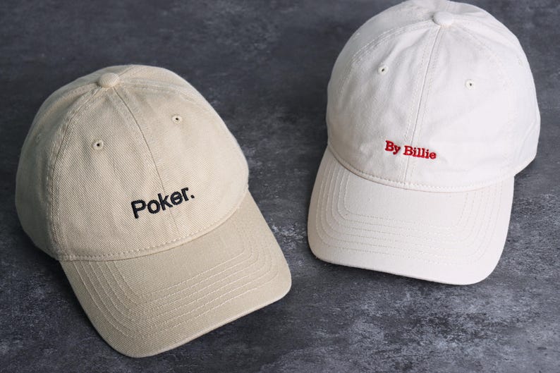 May include: Two baseball caps. One is tan with the word "Poker." embroidered in black. The other is white with "By Billie" embroidered in red. Both caps have a curved brim and adjustable strap.