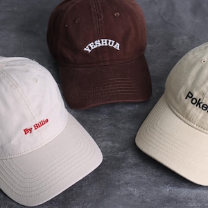 May include: Three baseball caps in different colors. One is brown with "Yeshua" embroidered in white. Another is beige with "By Billie" in red. The third is beige with "Poker." in black. All caps have a curved brim and adjustable strap.