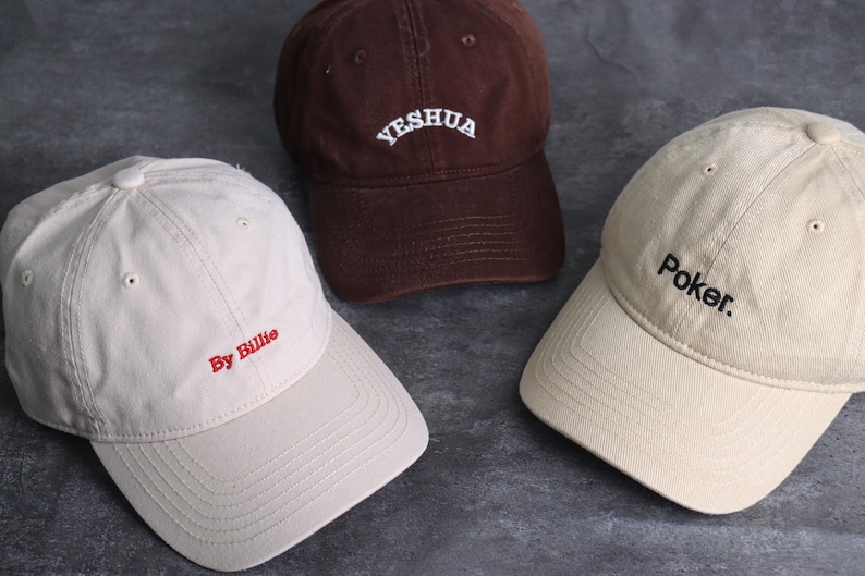 May include: Three baseball caps in different colors. One is brown with "Yeshua" embroidered in white. Another is beige with "By Billie" in red. The third is beige with "Poker." in black. All caps have a curved brim and adjustable strap.