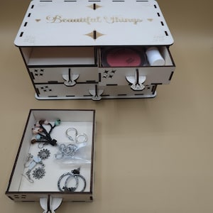 May include: A light-colored wooden jewelry box with three drawers and a top compartment. The top of the box has the words "Beautiful Things" engraved. The drawers contain makeup and jewelry.