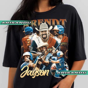 May include: Black t-shirt featuring a collage of images of a man in a cowboy hat, baseball uniform, and playing a guitar. The name "Jayson" is in gold script, and the word "ARENDT" is above the images.