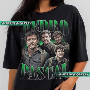 May include: Black t-shirt featuring a graphic of Pedro Pascal in various poses, with the name "PEDRO PASCAL" in green, stylized text. The shirt also includes the text "ARISANDU" in white. The design is a vintage style.
