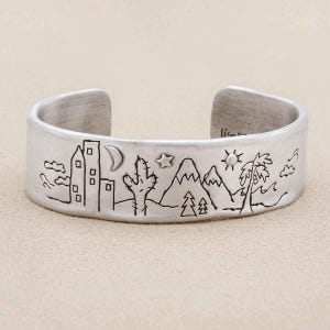 Puede incluir: Una pulsera de brazalete de metal plateado con un diseño de paisaje grabado. El grabado representa un horizonte urbano, una luna creciente, una estrella, un cactus, montañas nevadas, pinos, un sol y una palmera con líneas onduladas.