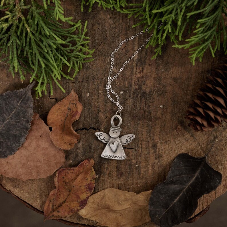 May include: A silver angel pendant necklace with a heart design. The pendant hangs on a silver chain, displayed on a rustic wooden surface with autumn leaves and evergreen branches. A charming piece of jewellery.