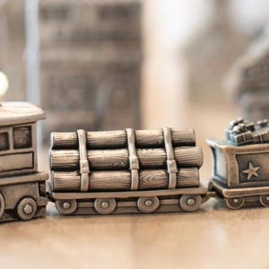 May include: A metallic silver-toned toy train set featuring a locomotive, a flatcar carrying a stack of cylindrical logs secured with straps, and a caboose decorated with a star emblem and small gift boxes on its roof, all with a weathered finish.