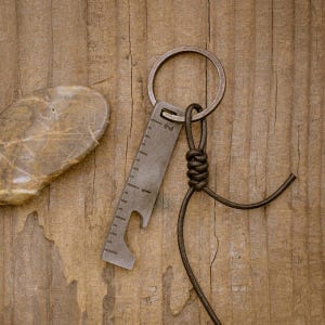 May include: A small, metal multi-tool keychain with a bottle opener and ruler markings. The keychain is attached to a dark brown leather cord. The background is a wooden surface.