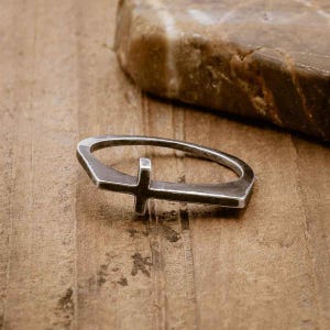 May include: A silver ring featuring a simple cross design. The ring is crafted from thick, textured metal and has a slightly rounded shape.