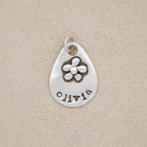 May include: Silver charm in the shape of a teardrop with a small flower design and the name "Olivia" engraved on it.