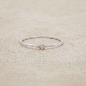May include: A delicate silver ring with a small, round, light pink gemstone set in the center. The ring has a thin band and a minimalist design, perfect for everyday wear. The ring is displayed on a neutral, textured surface.