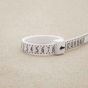 Puede incluir: Un medidor de anillos blanco con números negros del 2 al 17. El medidor de anillos está hecho de plástico y se utiliza para medir el tamaño de los dedos para anillos. Los números están impresos en una fuente clara y fácil de leer.