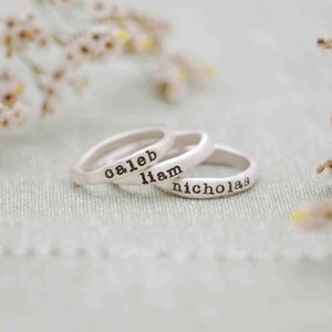 May include: Three silver rings with stamped lettering spelling out the names "caleb", "liam", and "nicholas". The rings are stacked on top of each other, with a soft-focus background of white and green.