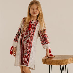 May include: A young girl wearing a white dress with red and black embroidered floral patterns. The dress has long sleeves and red tassels. She is standing next to a small wooden table with black metal legs, wearing black shoes.