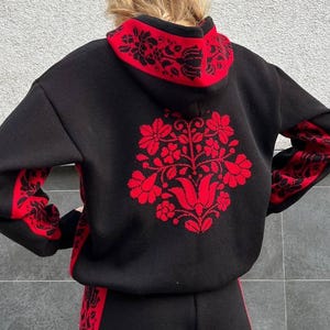 May include: Black hoodie and matching trousers set with red floral embroidery. The hoodie has a red-lined hood and floral design on the back and sleeves. The trousers have a red floral design along the sides. The outfit is made of a soft, warm material.