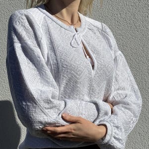 May include: White crocheted sweater with a tie at the neckline. The sweater has a textured pattern and long sleeves.