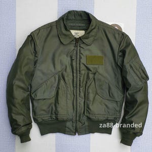 May include: An olive green bomber jacket with a zip-up front, two front pockets, and a patch on the chest. The jacket has ribbed cuffs and a collar. The sleeves have a pocket. The jacket is made of a smooth material, possibly nylon.