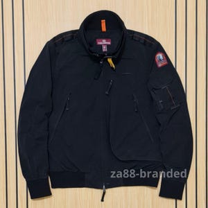Puede incluir: Una chaqueta bomber negra con cuello alto y lengüeta naranja, cierre frontal de cremallera con tirador amarillo, dos bolsillos inclinados en el pecho y un bolsillo utilitario en la manga izquierda con parche Parajumpers y costuras naranjas.