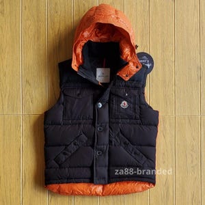 Puede incluir: Chaleco con capucha negro y naranja con diseño acolchado. El chaleco tiene cierre de botones en la parte delantera, dos bolsillos en el pecho y un parche con el logotipo. La capucha es de color naranja brillante y el chaleco tiene el texto "za88-branded".