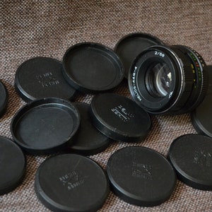 Puede incluir: Un objetivo de cámara Helios-44M-4 2/58 vintage con múltiples tapas de lente negras. El objetivo tiene un cuerpo negro y plateado con marcas coloridas. Las tapas de la lente están esparcidas sobre una superficie marrón texturizada.