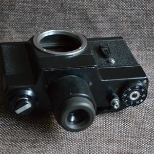 Könnte beinhalten: Eine schwarze Vintage-Filmkamera mit einem silbernen Objektivring und einem schwarzen Zifferblatt mit weißen Zahlen. Die Kamera hat einen schwarzen Lederriemen.