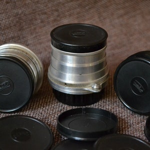 Op de afbeelding: Een set van zes zwarte camera lensdoppen met een zilveren lensdop in het midden. De lensdoppen hebben een reliëflogo aan de bovenkant.