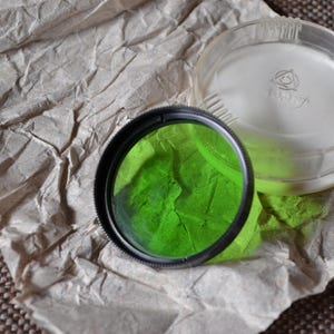 Könnte beinhalten: Ein grüner Filter mit schwarzem Rand liegt auf zerknittertem braunem Papier, neben einer transparenten Kunststoffhülle. Das Glas des Filters ist leuchtend grün. Die Hülle hat eine weiße Innenseite und ein Logo.