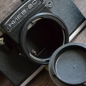May include: A black and silver vintage Kiev 60 camera with the lens cap removed. The camera body is black with a textured grip, and the lens mount is open, revealing the interior. The camera is positioned on a textured surface.
