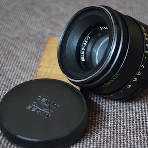 May include: A black camera lens with the text "HELIOS-44-2" printed on the side. The lens has a silver ring around the front and a black lens cap.