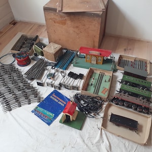 May include: A collection of vintage model train set components, including tracks, train cars, a station, and a control panel. The set includes a blue book with Russian text and a wooden storage box.