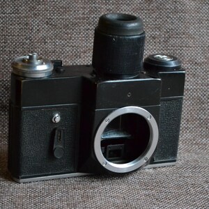 May include: A black and silver 35mm film camera with a lens attached. The camera has a viewfinder and a film advance lever.